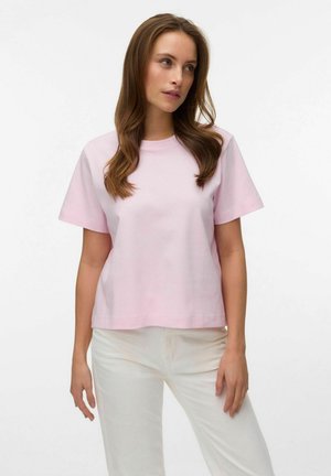 Kobieta o długich brązowych włosach, ubrana w jasnoróżową koszulkę z krótkim rękawem i białe spodnie, stojąca na tle jednolitej białej ściany.