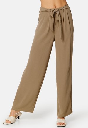 Beżowe szerokie spodnie z wysokim stanem z przodu związane paskiem, noszone z białymi sandałami na obcasie z odkrytymi palcami, widoczne od talii do stóp.