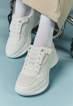 Baskets blanches épaisses avec des détails en mesh et cuir portées avec des chaussettes blanches et un pantalon beige, sur un sol bleu clair à côté d'un fauteuil roulant.