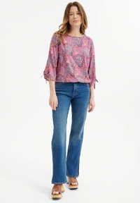 Rosa Bluse mit Paisleymuster und Dreiviertelärmeln, kombiniert mit blauen Schlagjeans und Holz-Plateau-Sandalen.
