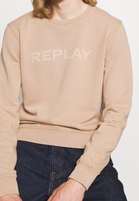 Felpa beige con scollatura a girocollo e logo "REPLAY" leggermente in rilievo al centro. Il tessuto appare morbido con polsini e orlo a costine.