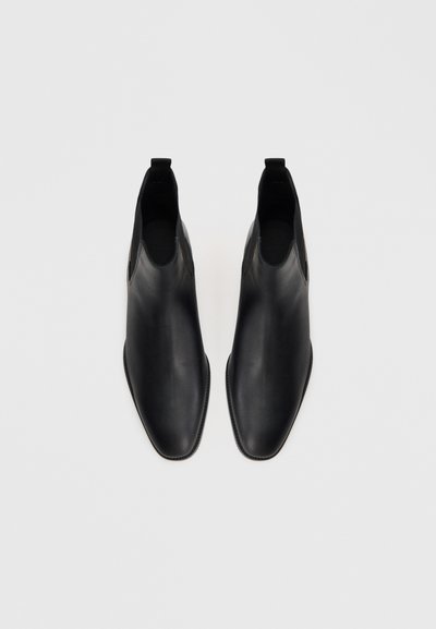 Pár fekete bőr Chelsea csizma elasztikus oldalpanelekkel, lekerekített négyzet alakú orral és húzófülekkel, fehér háttéren.