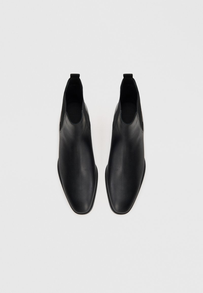 Paire de bottes Chelsea en cuir noir avec panneaux élastiques sur les côtés, bouts carrés arrondis et tirettes sur fond blanc.