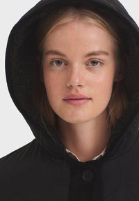 Veste matelassée noire avec capuche doublée en tissu texturé, présentant une fermeture à boutons et de subtils détails de couture.
