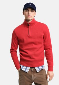 Roter Reißverschluss-Pullover mit hohem Kragen, mit gerippten Bündchen und Saum. Trägt man über einem hellblau-weiß gestreiften Hemd.