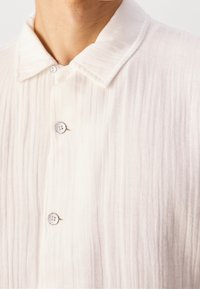 Gros plan d’une personne portant une chemise blanche texturée à boutons, avec un col classique et trois boutons visibles fermés.