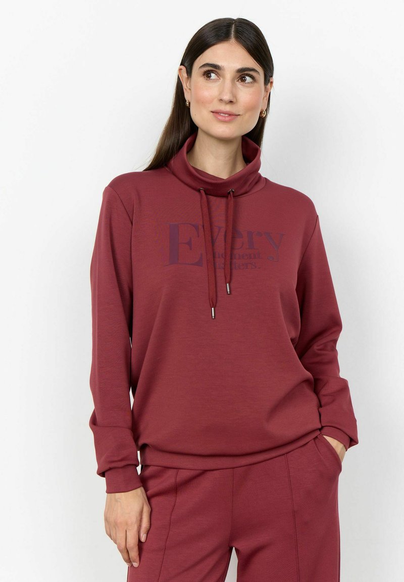 Femme aux longs cheveux foncés portant des boucles d'oreilles créoles en or et un ensemble sweat-shirt et pantalon bordeaux, debout les mains dans les poches, regardant vers la droite.