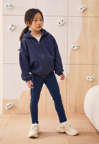 Temno modra zip-up kapuca s sprednjimi žepi, v kombinaciji z temno modrimi leggingsi in bež športnimi copati. Ima sproščen kroj in mehak material.