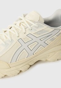 Beige spordijalatsid, millel on võrkpealne osa, hallid detailid, tekstureeritud põhi ja kontrastne logo. Paelad ja pehme krae suurendavad mugavust.