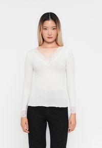 Jeune femme avec de longs cheveux ombrés portant un haut blanc à manches longues avec des dentelles et un pantalon noir, se tenant devant un fond uni.