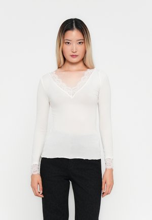 Jeune femme avec de longs cheveux ombrés portant un haut blanc à manches longues avec des dentelles et un pantalon noir, se tenant devant un fond uni.