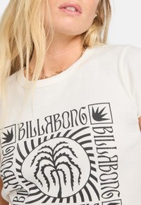 T-shirt a righe color crema con un design grafico nero che presenta un motivo vegetale e schemi variabili. Il collo è rotondo e aderente.