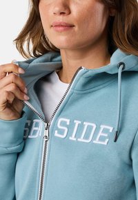 Lichtblauwe zip-up hoodie van zachte stof met witte geborduurde tekst "SEASIDE", capuchon met trekkoorden en zilveren ritsdetail.