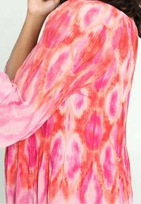 Persona che indossa un capo a maniche lunghe in tie-dye rosa e rosso con texture a coste, braccio sollevato vicino alla testa su uno sfondo chiaro.