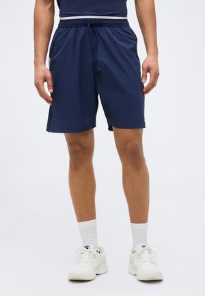 Egy férfi sötétkék sportnadrágot, fehér bokazoknit és fehér tornacipőt visel, és egy egyszerű világos háttér előtt áll.