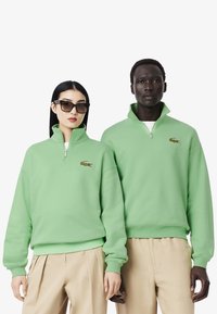 Sudadera verde con cremallera hecha de tejido suave, con cuello acanalado, logo bordado y corte relajado. Llevada con pantalones beiges.