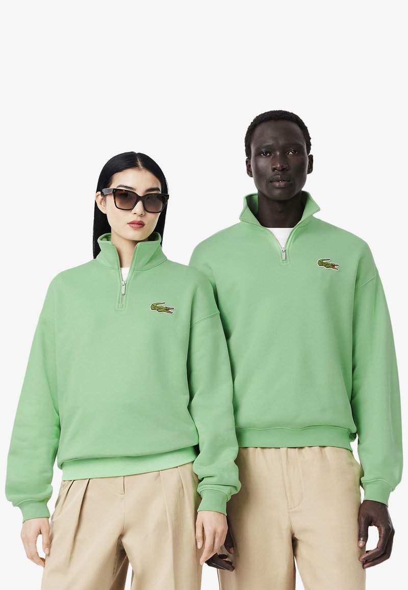 Sudadera verde con cremallera hecha de tejido suave, con cuello acanalado, logo bordado y corte relajado. Llevada con pantalones beiges.