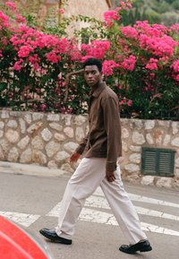 Man in brown shirt and white pants walking across a street with stone wall and vibrant pink flowers in the background.
