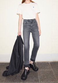 T-shirt beige clair avec un accent bleu, jean gris taille haute, sabots noirs, et une veste en jean noire drapée sur le bras contre un mur clair.