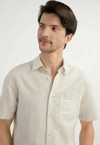 Jasno-beżowa koszula z krótkim rękawem z lnu, z kołnierzykiem zapinanym na guziki, kieszenią z przodu oraz subtelną teksturą w jednolitym wzorze.