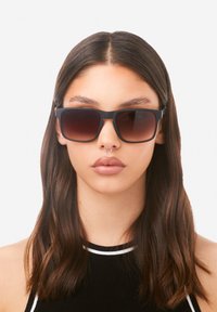 Musta suorakaiteen muotoinen aurinkolasit, joissa on liukuvärjätyt tummat linssit, mattapintainen rakenne ja paksut kehykset. Käytössä henkilöllä, jolla on pitkä, tumma tukka.