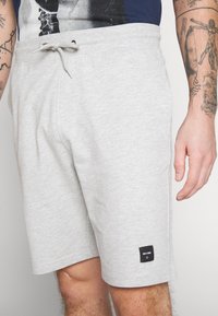 Pantalones cortos de algodón gris claro con cintura elástica y cordón. Presentan bolsillos laterales y un pequeño parche negro en el dobladillo.