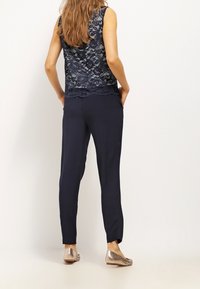 Haut sans manches en dentelle marine avec ourlet festonné, associé à un pantalon foncé. Personne portant des ballerines métalliques, debout les mains dans les poches.