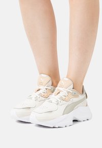 Baskets athlétiques blanches et beiges avec une tige en mesh texturé, une languette rembourrée et une semelle blanche épaisse. Comprend des logos subtils et des détails d'accent.