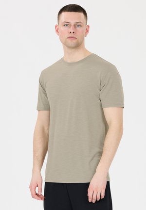 Mand med kort hår iført en beige kortærmet T-shirt og sorte bukser, stående mod en ensfarvet hvid baggrund.