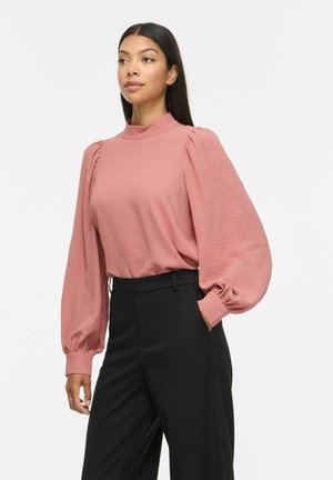 Vrouw draagt een roze blouse met lange pofmouwen en zwarte hoge taille broek, staand met één hand in de zak.