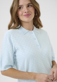 Helles blaues Poloshirt mit kurzen Ärmeln aus weichem Stoff, versehen mit einem strukturierten Rautenmuster, einer dreiknöpfigen Knopfleiste und einem klassischen Kragen.
