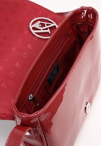 Borsa a mano in pelle verniciata rossa aperta per mostrare la tasca con zip interna e la pattina foderata in tessuto con loghi ripetuti "AJ" e triangoli.