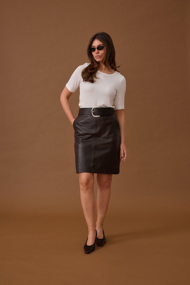 Femme portant des lunettes de soleil noires, un haut blanc à manches courtes, une jupe en cuir noire avec une large ceinture et des talons noirs, debout devant un fond marron.