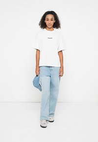 White cotton t-shirt with short sleeves and a black printed logo. Paired with light blue denim jeans and grey sneakers. Light blue bag on arm.