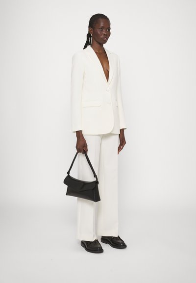 Costume blanc avec un blazer à col en V profond et un pantalon large. Le modèle tient un sac à main en cuir noir. Texture lisse, design ajusté, style minimaliste.