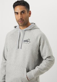 Grå sweatshirt med hætte, med lomme foran og raglanærmer. Broderet grafik af en lastbil med en gravemaskine som accent.