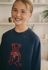 Merkkihuppari, jossa on punaisella brodeerattu karhu, joka pitää nauhaa. Pehmeä kangas, rento malli ja raglan-hihat.