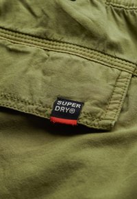 Olivengroen stof met een gestructureerd oppervlak, voorzien van een zwart etiket met witte tekst "SUPER DRY" en een rode accent eronder.