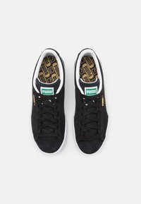 Baskets en suede noir avec semelles en caoutchouc blanc, logo Puma vert et blanc sur la languette, et lacets noirs. Semelle intérieure texturée avec motif de logo.