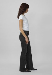 Femeie în profil, îmbrăcată cu o bluză albă cu mânecă scurtă, blugi negri evazați cu talie înaltă și pantofi negri cu vârful ascuțit, pe un fundal gri.