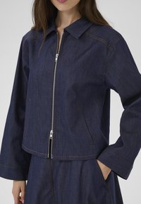 Jeansowa kurtka w ciemnoniebieskim kolorze z zamkiem błyskawicznym z przodu, spiczastym kołnierzem i dwoma bocznymi kieszeniami. Posiada kontrastowe żółte przeszycia i luźny krój.