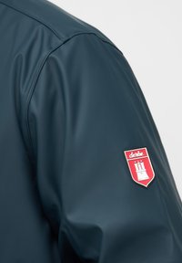 Giacca impermeabile di un blu scuro che presenta una texture liscia, dettagli sulle cuciture delle spalle e un logo a forma di scudo rosso sulla manica destra.