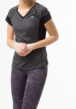 Kvinde iført mørkegrå kortærmet træningstrøje og mønstrede lilla leggings, stående med hænderne sammen foran.