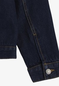 Manica di giacca in denim blu scuro con cuciture dorate e dettaglio di un bottone metallico al polsino. Texture liscia con orlo piegato.