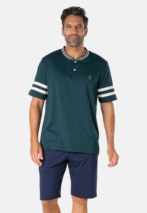 Homme aux cheveux courts et foncés portant une chemise à manches courtes vert foncé avec des rayures blanches sur les manches et un short bleu marine.
