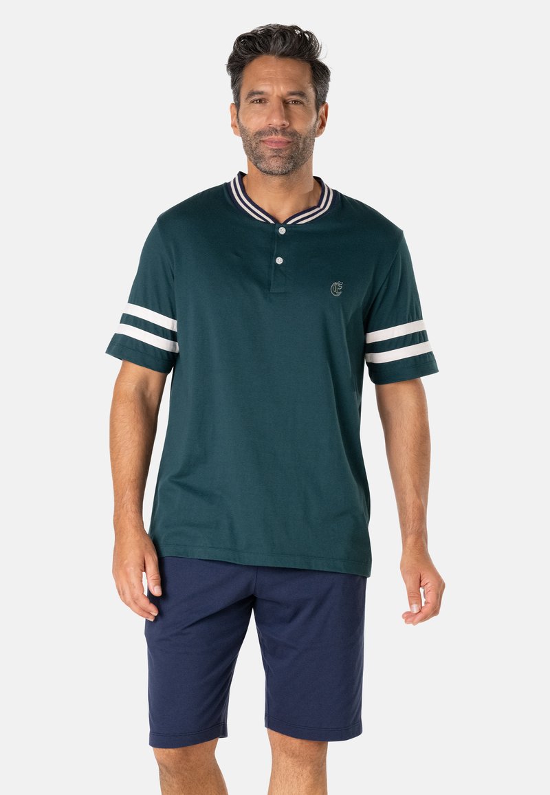 Homme aux cheveux courts et foncés portant une chemise à manches courtes vert foncé avec des rayures blanches sur les manches et un short bleu marine.