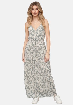 Kvinde iført enærmet, grå maxikjole med hvidt blomstret print kombineret med hvide sneakers, stående mod en ensfarvet baggrund.