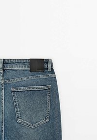 Farkut keskisinisessä pesussa, joissa on musta nahkatagi takavyötäröllä ja yksi takatasku, jossa on ompelusdetaili.