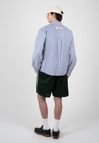 Hombre con gorra beige, camisa holgada de rayas verticales azules y rosas, pantalones cortos verde oscuro con rayas laterales, calcetines blancos y zapatos negros, de espaldas.
