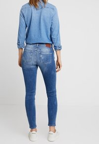 Jeans de mezclilla con un degradado azul, corte slim, diseño de cinco bolsillos y una parche de cuero marrón en la parte trasera. Combinados con zapatillas blancas.